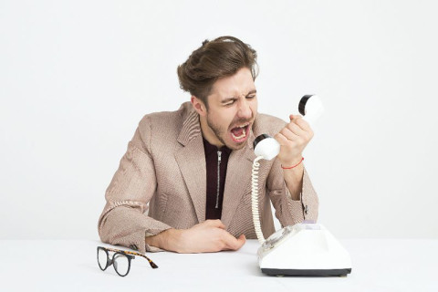 Man in tan jacket and black shirt yelling into a white phone. Eye glasses sitting on desk near the phone.