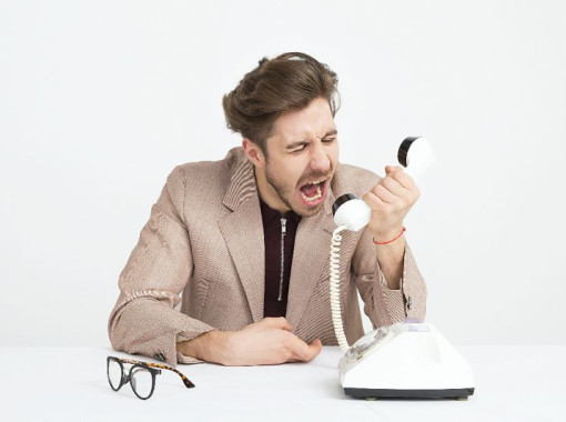 Man in tan jacket and black shirt yelling into a white phone. Eye glasses sitting on desk near the phone.